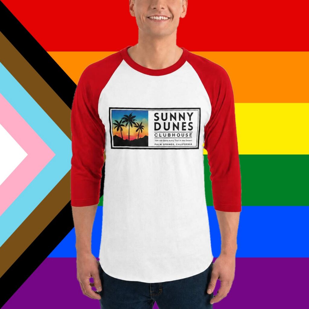 Person wearing a white and red 3/4-sleeve raglan shirt with the Sunny Dunes Clubhouse logo, in front of a Progress Pride flag